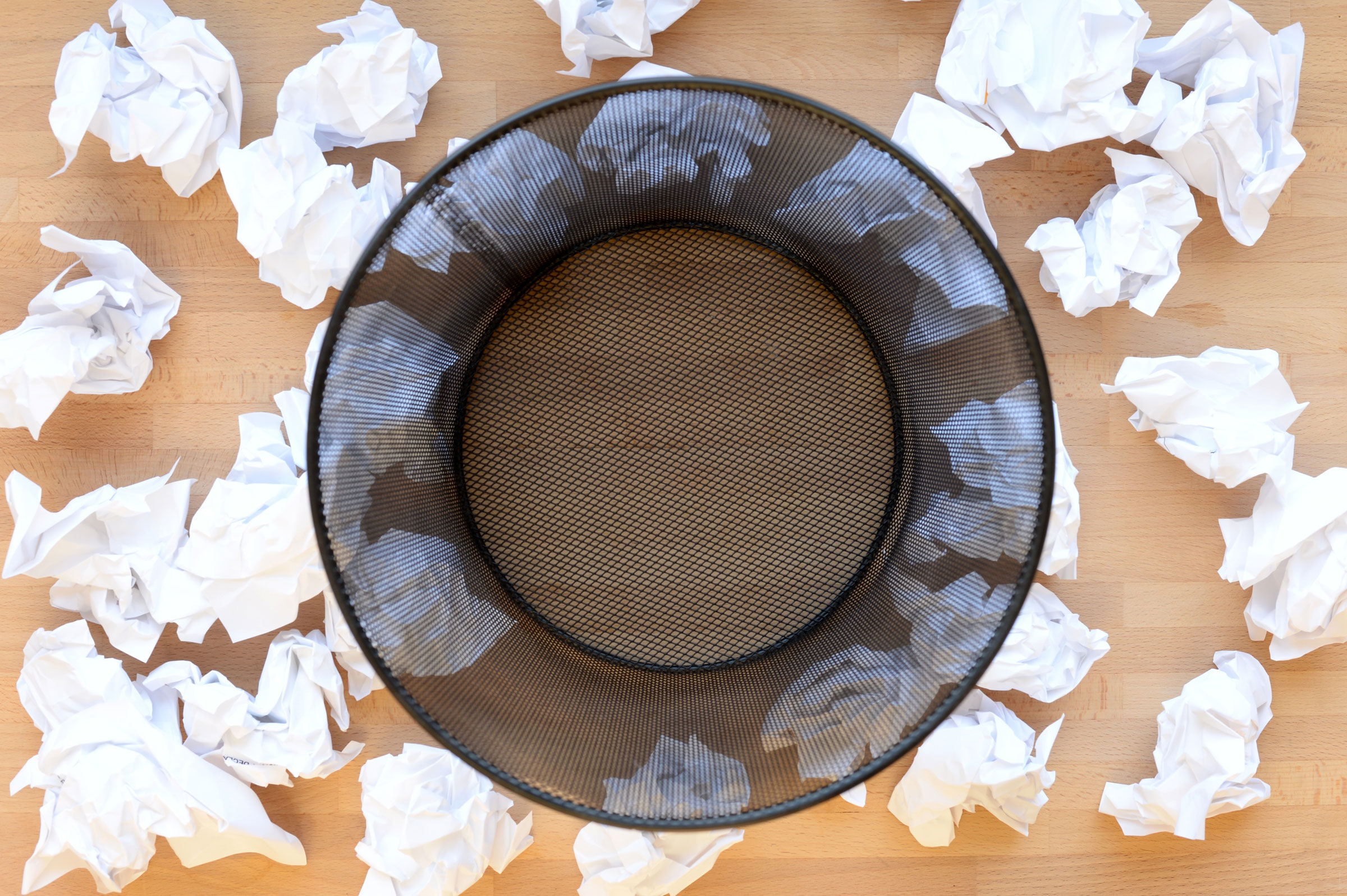 Overhead view of a black mesh wastebasket surrounded by crumpled sheets of white paper on a wooden floor.